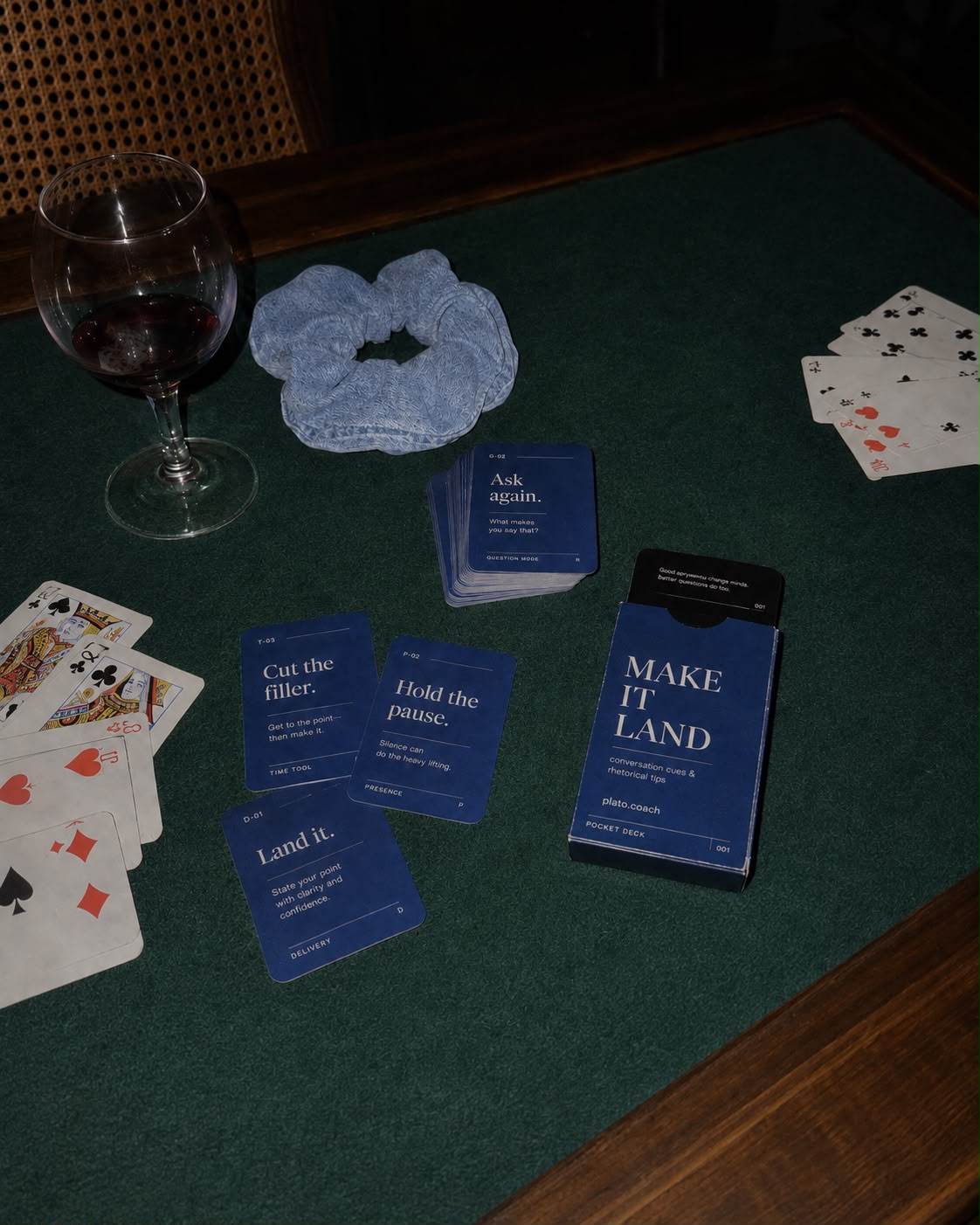 Blue Make It Land pocket deck and conversation cue cards spread on a green card table beside playing cards and a glass of red wine