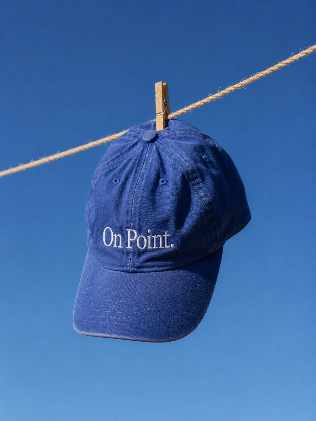 Navy Plato cap embroidered with the words On Point. hanging from a washing line against a blue sky